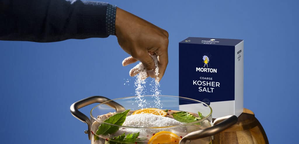 Hand sprinkling Morton coarse kosher salt into a glass bowl with marinated ingredients, showcasing the product packaging against a blue background, highlighting culinary use and flavor enhancement.