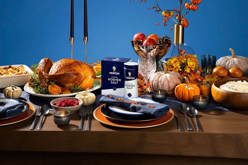 Thanksgiving table setting featuring a roasted turkey garnished with herbs and oranges, alongside side dishes like mashed potatoes and cranberry sauce. Morton's coarse kosher salt is prominently displayed, with decorative pumpkins and autumn foliage enhancing the festive atmosphere.