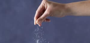Hand sprinkling salt against a blurred gray background, illustrating the culinary use of seasoning in cooking.