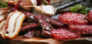 Assorted cured meats including bacon, salami, and jerky, garnished with garlic and herbs, displayed on a wooden cutting board. Ideal for charcuterie boards or gourmet appetizers.