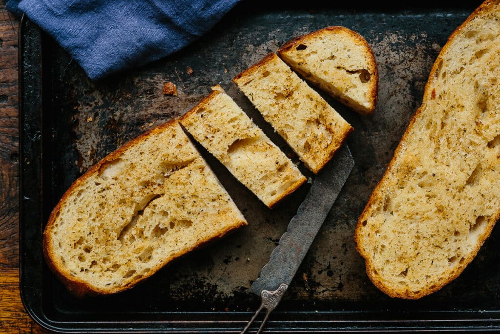 Sliced homemade bread on a dark baking tray with a serrated knife, showcasing a golden-brown crust and soft, airy interior. A blue linen cloth is partially visible in the background, adding a rustic touch to the scene.