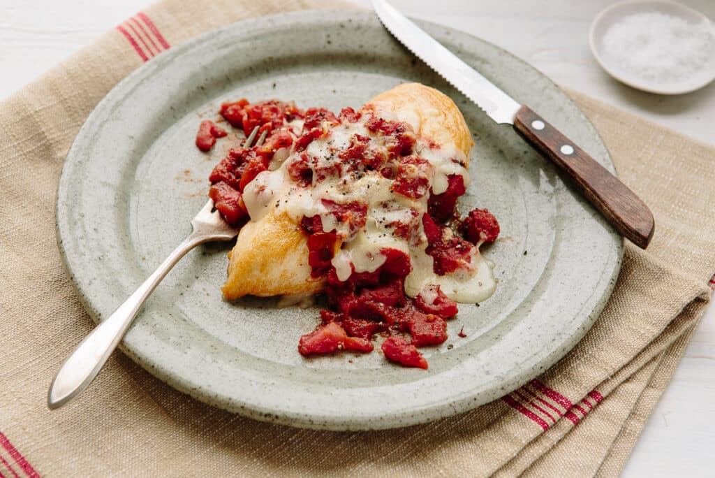 Delicious chicken topped with savory diced tomatoes and melted cheese, served on a rustic plate with a fork and knife, against a textured linen backdrop.