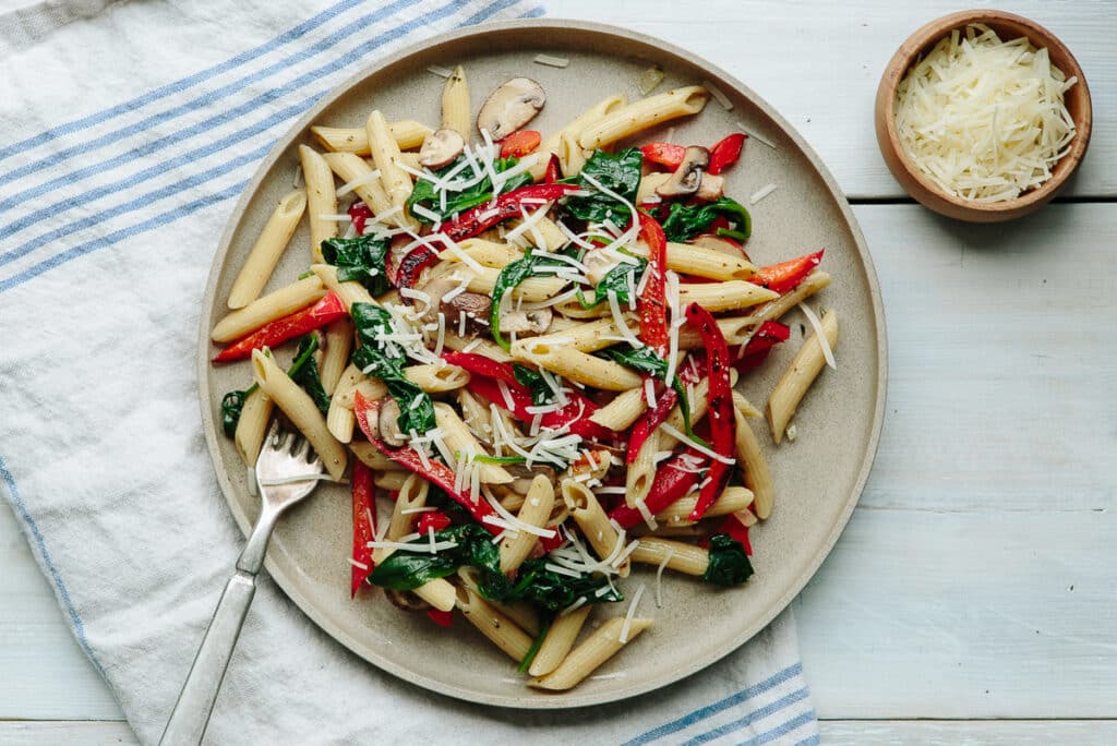 Penne pasta dish featuring sautéed spinach, red bell peppers, and mushrooms, topped with grated cheese, served on a beige plate with a fork, alongside a small bowl of shredded cheese, against a light wooden background.