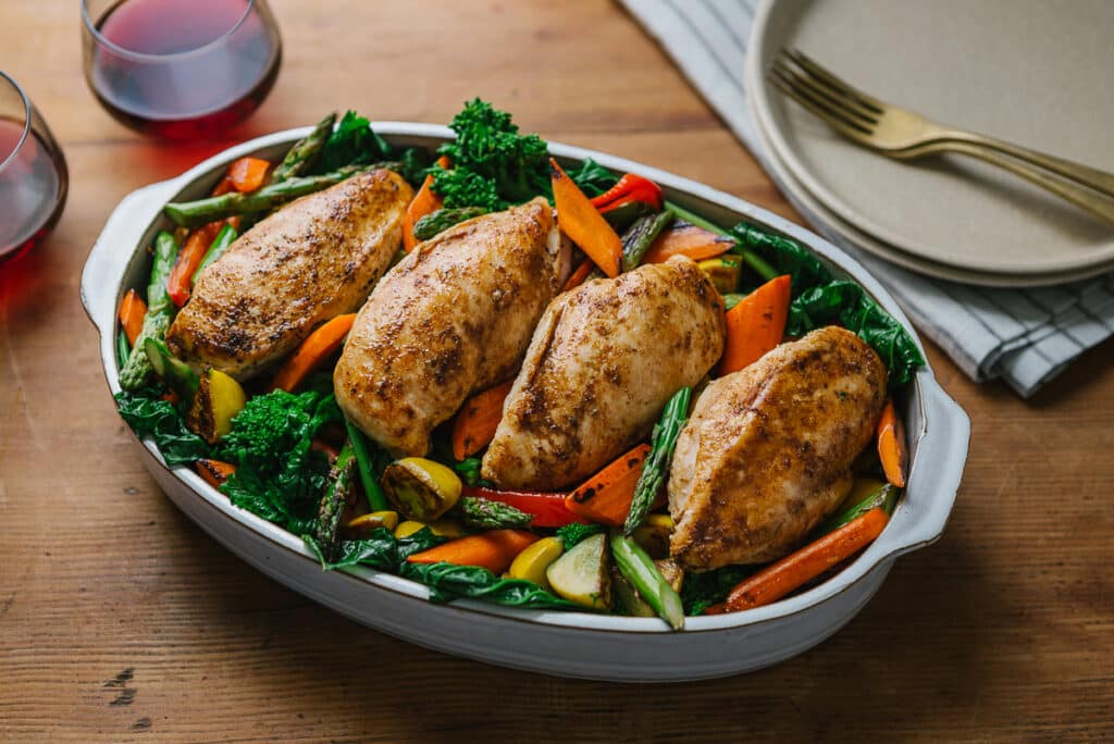 Baked chicken breasts served on a bed of colorful roasted vegetables, including carrots, asparagus, and leafy greens, with two glasses of red wine in the background.