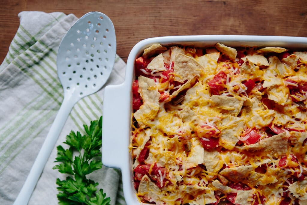 Baked tortilla casserole topped with melted cheese and crispy tortilla pieces, served with a slotted spoon and fresh parsley on a wooden table.