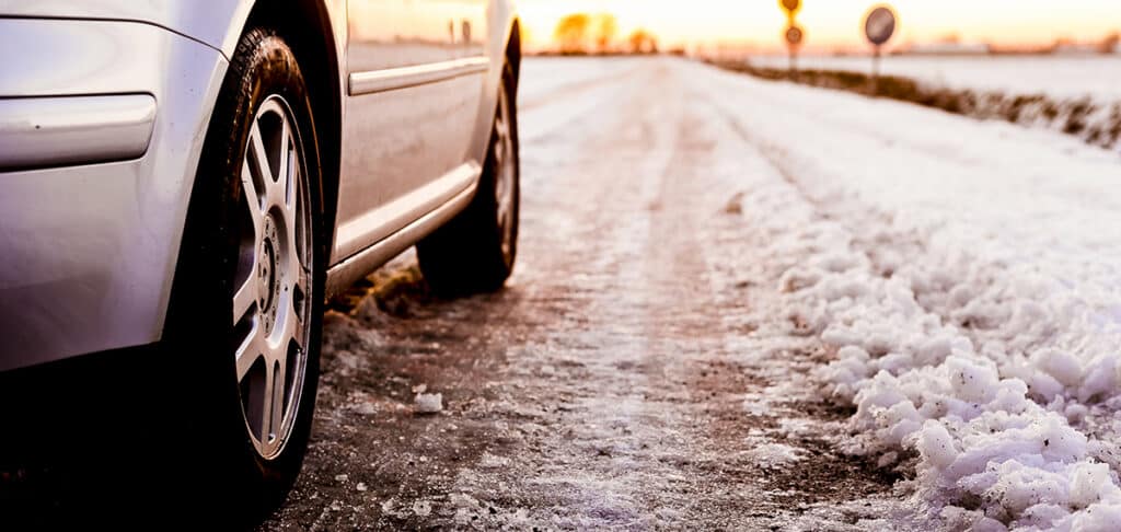 A silver car parked on a snow-covered road during sunset, showcasing icy conditions and winter travel challenges.