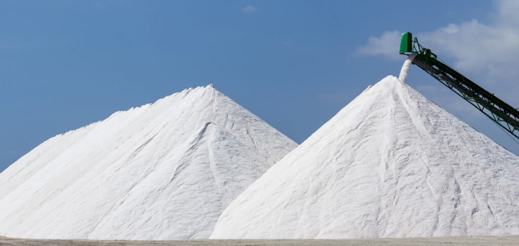 Piles of white salt with a conveyor belt transporting salt in a bright blue sky setting, showcasing salt production and storage.