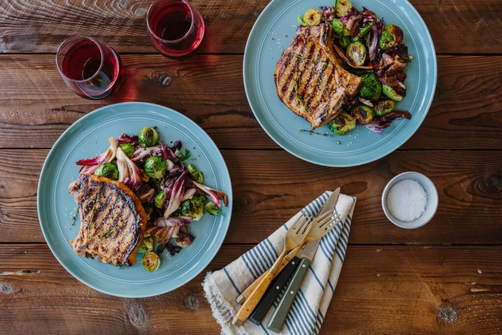 Grilled pork chops served with roasted Brussels sprouts and mixed greens on light blue plates, accompanied by two glasses of red wine and a small bowl of salt on a wooden table.