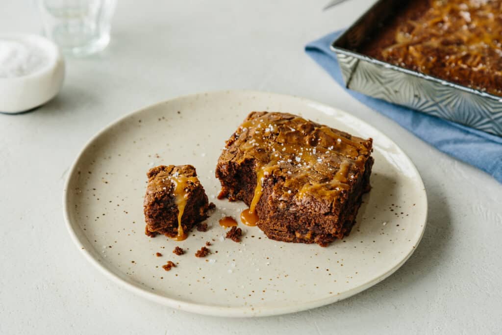 Delicious caramel brownie on a speckled plate, with a small piece cut out, showcasing a gooey caramel drizzle and a sprinkle of sea salt. A baking dish with more brownies is visible in the background.