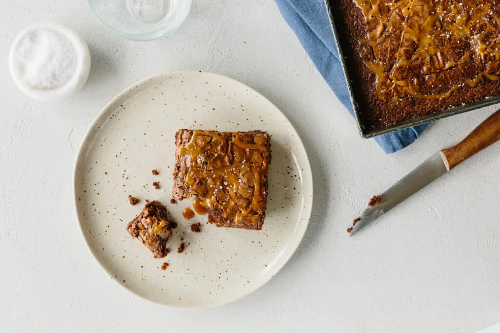 Delicious chocolate brownie squares topped with caramel sauce and sea salt, served on a speckled plate with a small piece cut out, accompanied by a glass of water and a bowl of salt, against a light textured background.