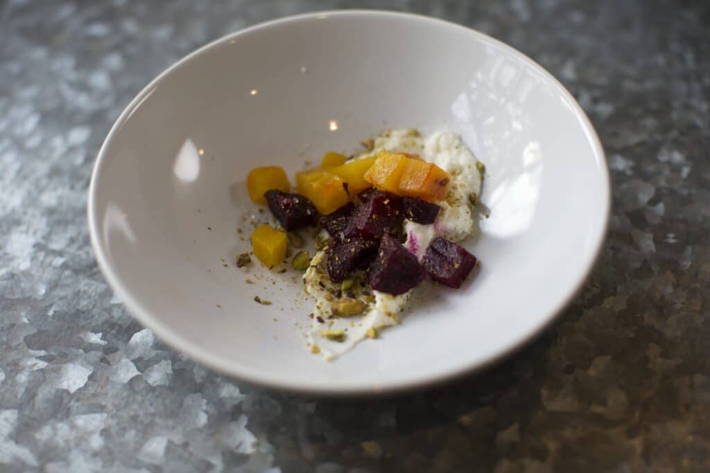 Colorful roasted beets and squash served on a bed of creamy yogurt, garnished with crushed pistachios and spices, presented in a white bowl on a textured metal surface.