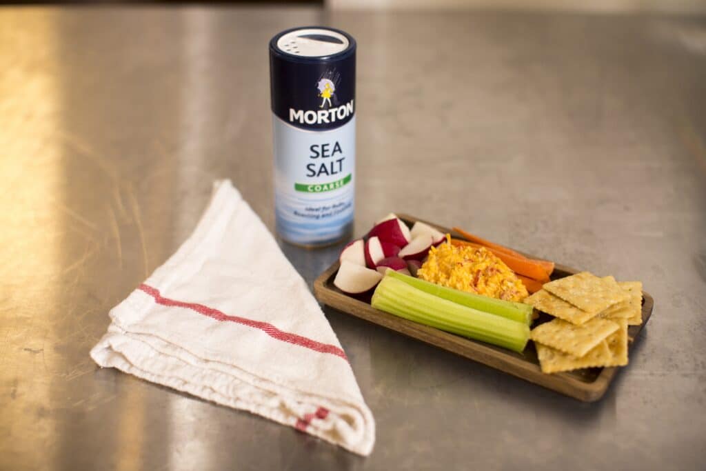 A wooden platter featuring a colorful assortment of fresh vegetables, including celery, carrots, and radishes, accompanied by a creamy dip and crackers. A canister of Morton coarse sea salt and a folded kitchen towel are also visible in the scene, set against a stainless steel surface.