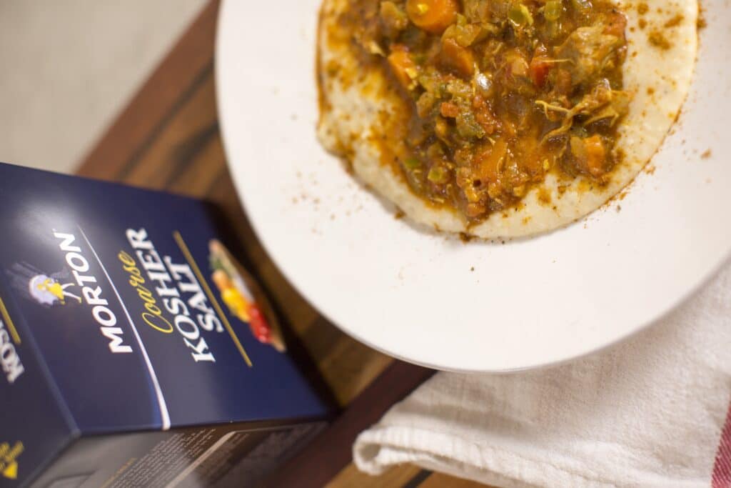 Bowl of creamy grits topped with a savory vegetable and meat stew, accompanied by a box of Morton coarse kosher salt on a wooden surface.