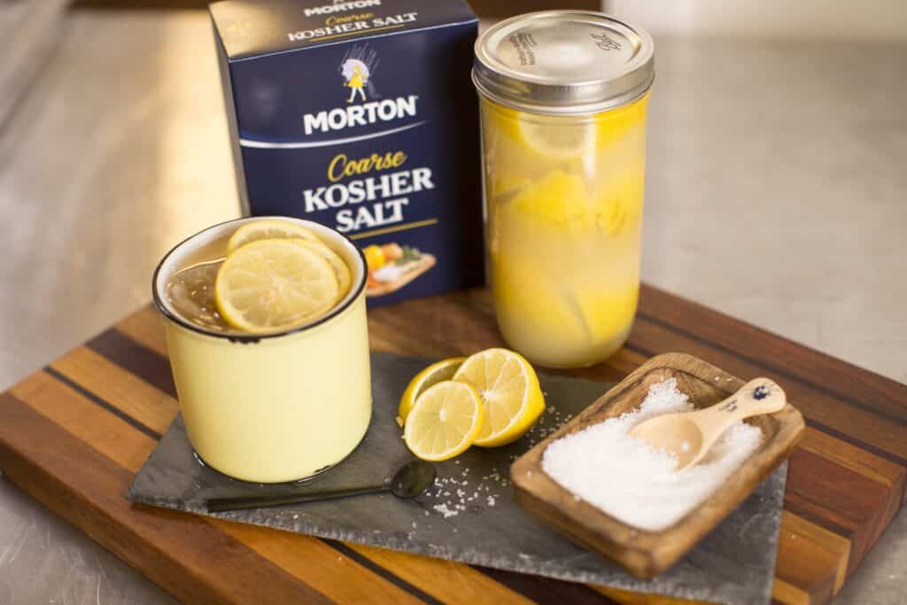 Image of a yellow cup filled with a lemon drink, garnished with lemon slices, alongside a jar of preserved lemons, a container of Morton coarse kosher salt, and fresh lemon halves on a wooden cutting board. The scene highlights ingredients for cooking and beverage preparation.