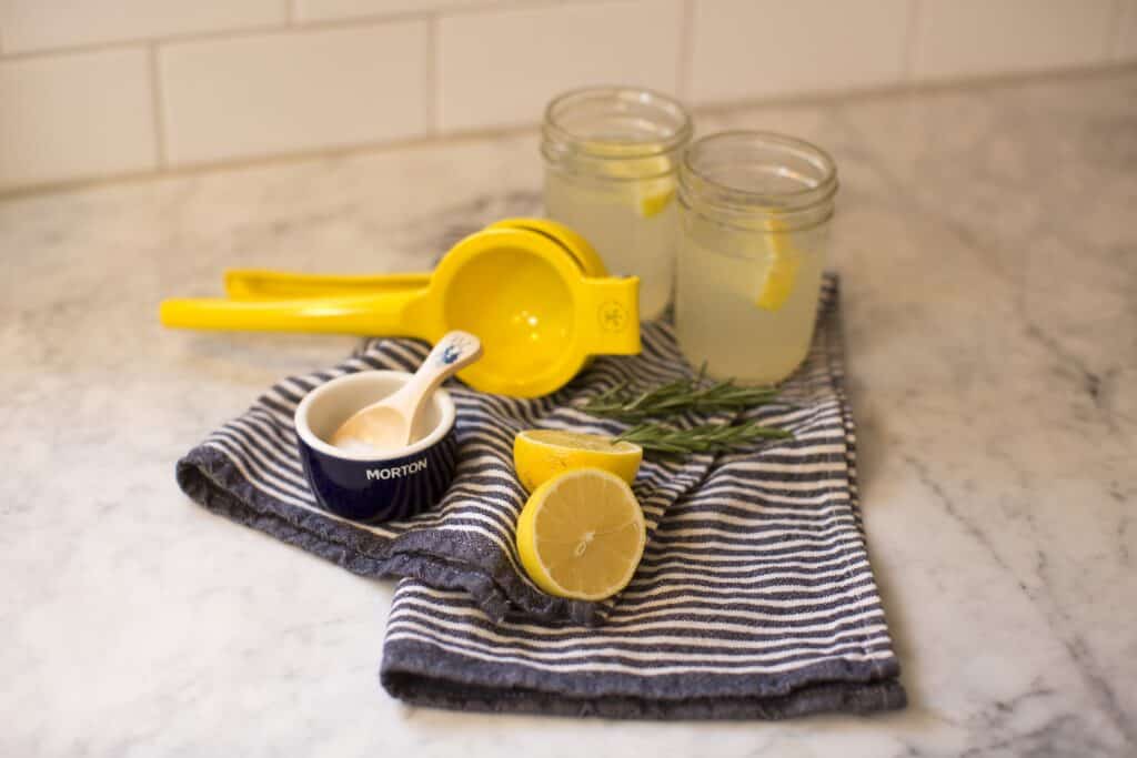 Freshly squeezed lemonade preparation with yellow citrus juicer, lemon halves, salt, and rosemary on a striped kitchen towel. Two glasses filled with lemonade and garnished with lemon slices are visible in the background.