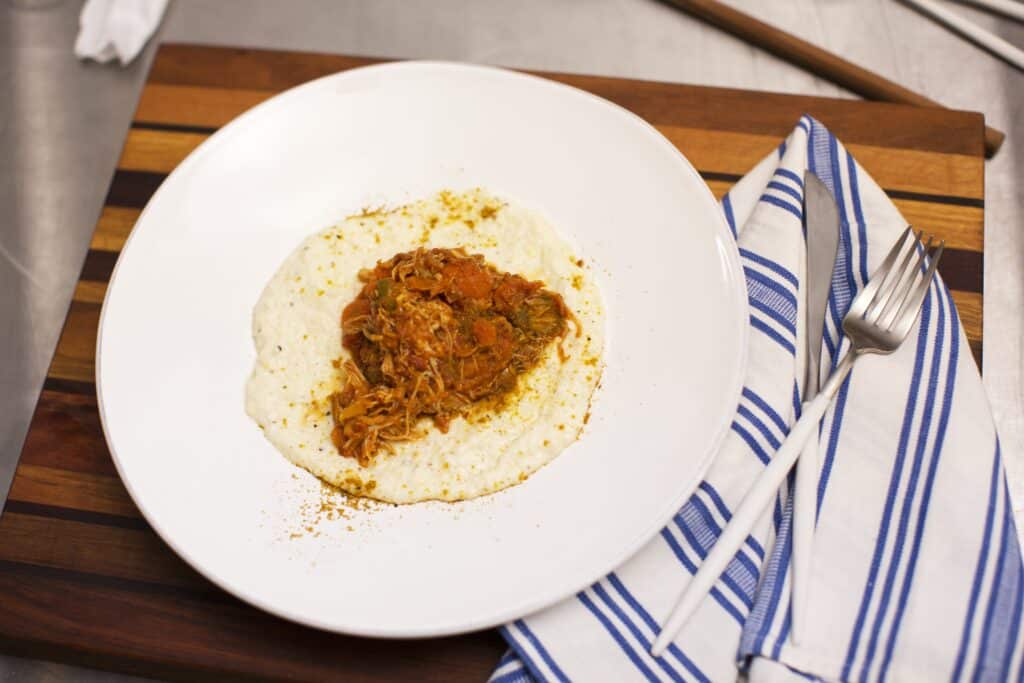 Plate of creamy grits topped with savory pulled chicken and tomato sauce, garnished with spices, served on a wooden cutting board with a striped napkin and fork.