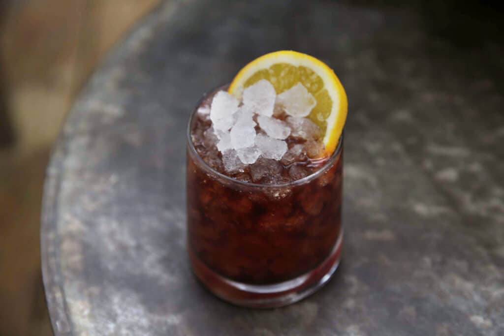 Refreshing cocktail in a glass filled with crushed ice, garnished with a slice of lemon, served on a rustic metal table. Perfect for summer gatherings or casual sipping.