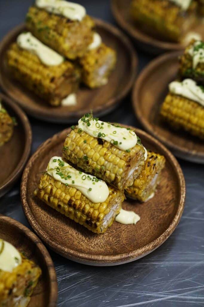 Grilled corn on the cob topped with creamy sauce and chives, served on wooden plates, showcasing a delicious appetizer or side dish.