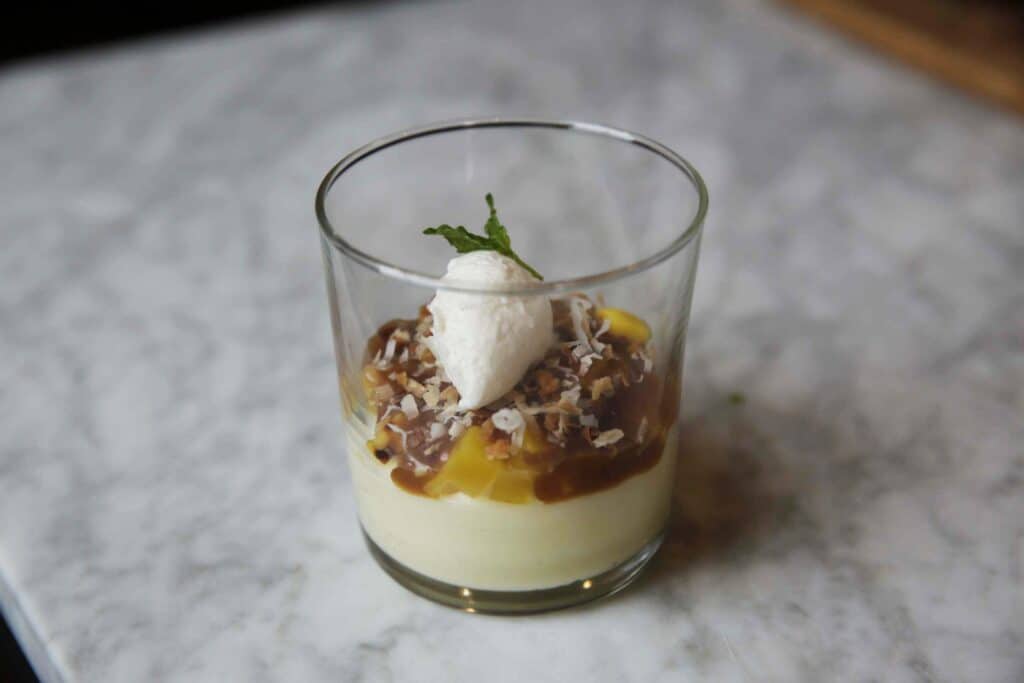 Delicious dessert in a glass featuring layers of creamy vanilla pudding, caramel sauce, fresh mango, whipped cream, and sprinkled with toasted coconut flakes, garnished with a mint leaf, served on a marble countertop.
