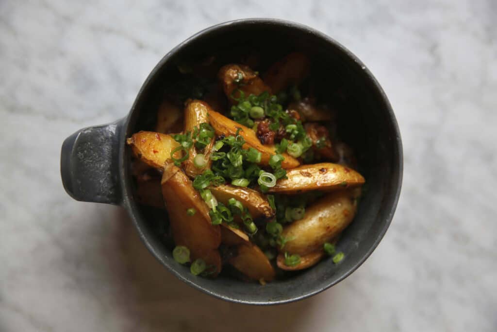 Deliciously cooked golden potatoes garnished with fresh green onions in a dark bowl, set against a light marble background, perfect for a hearty side dish or appetizer.
