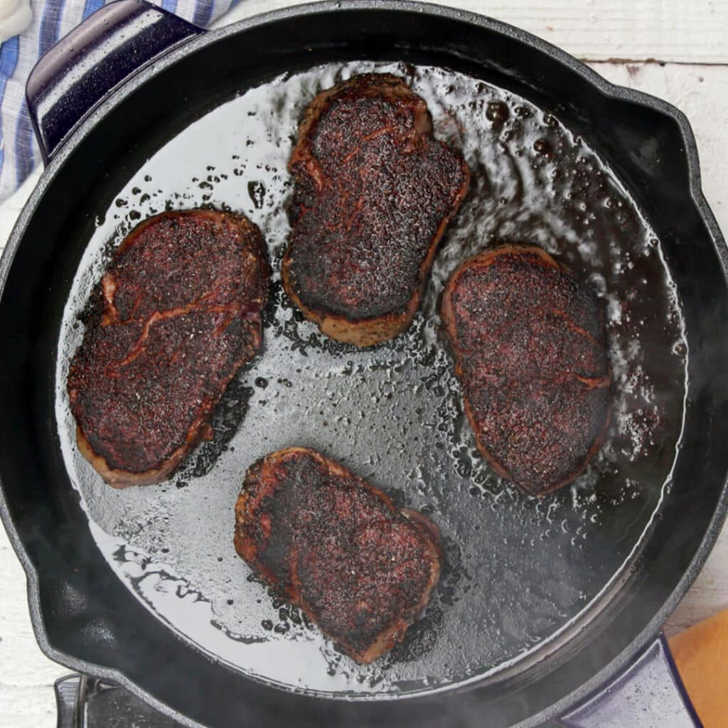 Four perfectly seared steaks cooking in a cast iron skillet, surrounded by sizzling oil and steam, showcasing a rich, dark crust.