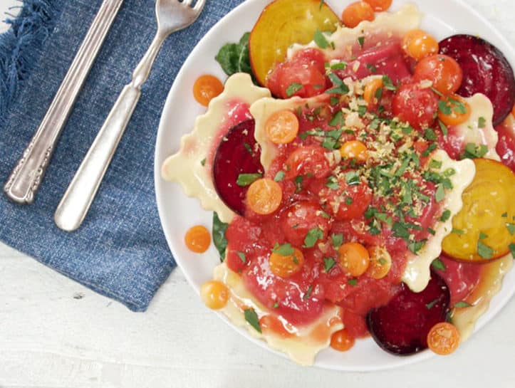 Colorful plate of ravioli topped with fresh tomatoes, beets, and herbs, served on a blue linen napkin with vintage silverware, showcasing a vibrant and appetizing dish.