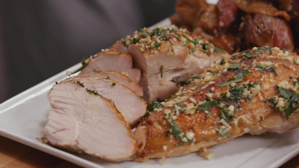 Sliced roasted turkey breast garnished with herbs and garlic, served on a white platter alongside crispy skin turkey pieces. Ideal for holiday meals and festive gatherings.
