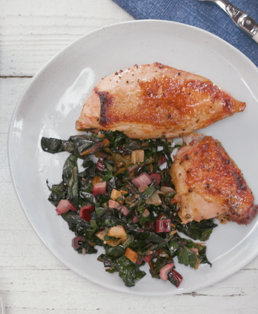 Grilled chicken breasts served on a white plate with a colorful sauté of Swiss chard, onions, and peppers. The dish is set against a rustic wooden background, highlighting a healthy and vibrant meal option.
