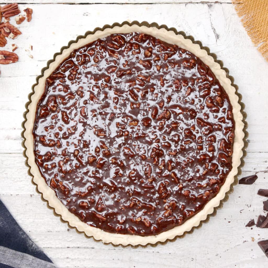 Chocolate pecan pie in a tart shell, featuring a glossy chocolate filling topped with chopped pecans, placed on a rustic wooden surface with chocolate pieces nearby.