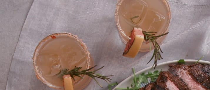 Two refreshing cocktails garnished with lemon and rosemary beside a plate of grilled steak, showcasing a delicious outdoor dining setup.
