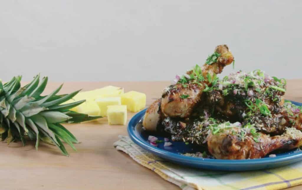 Grilled chicken drumsticks garnished with green onions and red onions, served on a blue plate alongside fresh pineapple chunks on a wooden table.
