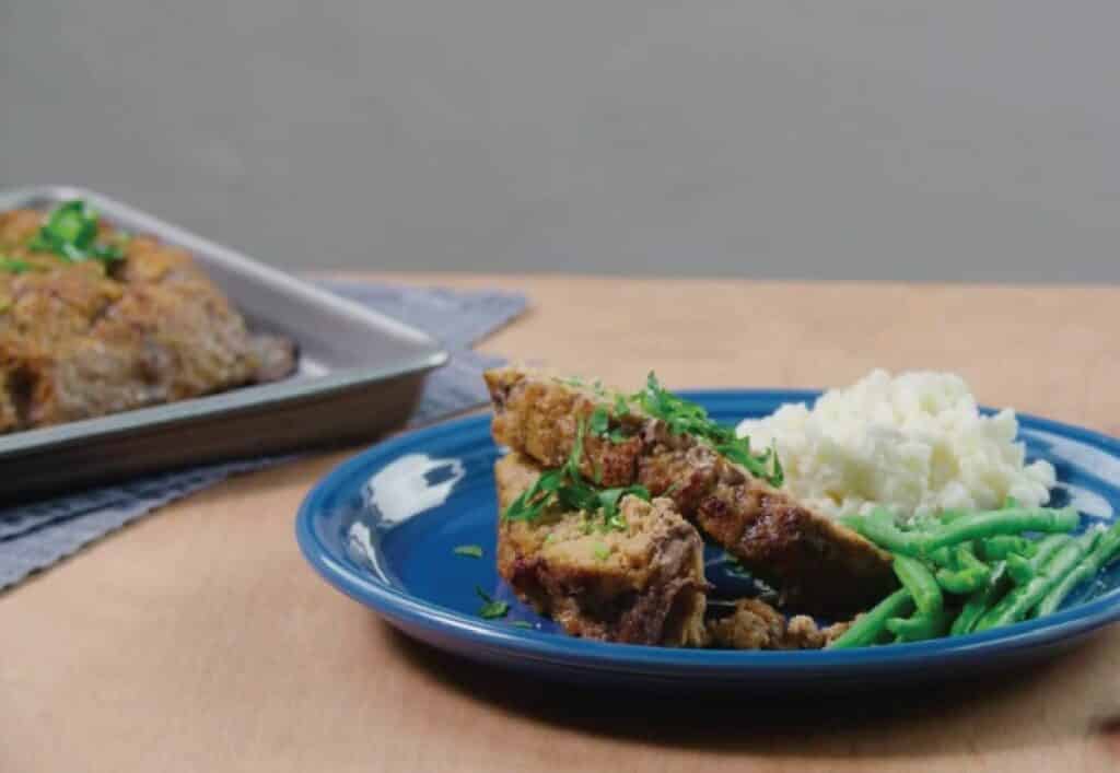 Plate of meatloaf slices garnished with parsley, served alongside creamy mashed potatoes and steamed green beans, with a baking tray of meatloaf in the background.
