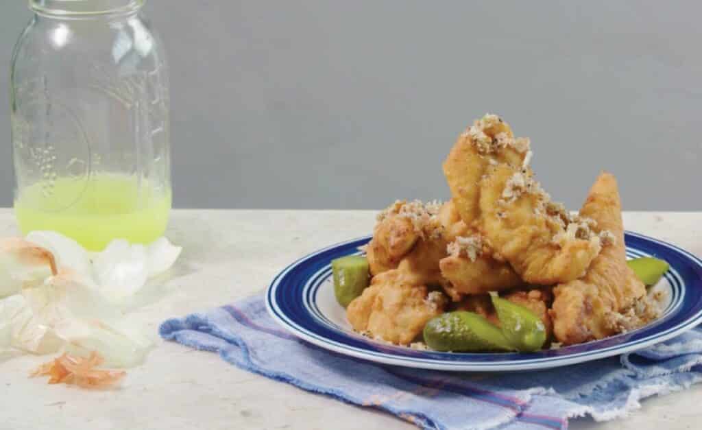 Plate of crispy fried fish garnished with shredded coconut, served with pickles on a blue and white patterned dish, accompanied by a jar of yellow beverage and a decorative cloth.