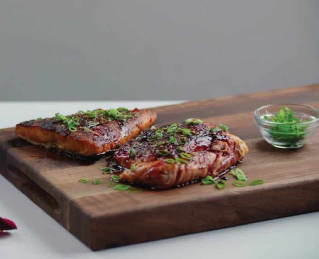 Grilled salmon fillets garnished with chopped green onions, served on a wooden cutting board with a small bowl of fresh herbs.