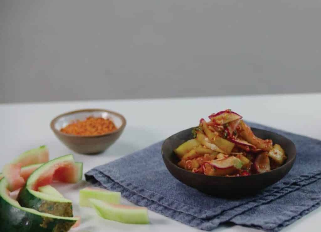 Bowl of spicy watermelon salad with colorful vegetables and spices, served on a blue cloth, with a side dish of chili powder and watermelon slices.