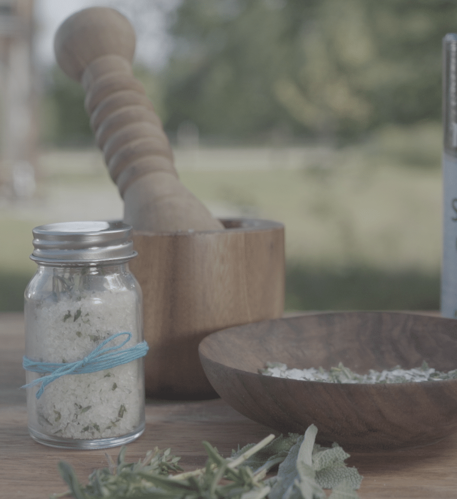 Glass jar of herb-infused salt with a blue twine bow, wooden mortar and pestle in the background, and a wooden bowl with salt and herbs on a rustic table, surrounded by fresh herbs outdoors.
