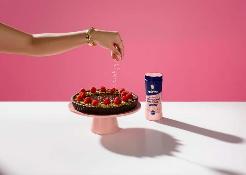 A hand sprinkles coarse Himalayan pink salt over a chocolate tart decorated with raspberries and pistachios, with a pink background and a container of Morton Himalayan pink salt prominently displayed beside the dessert.