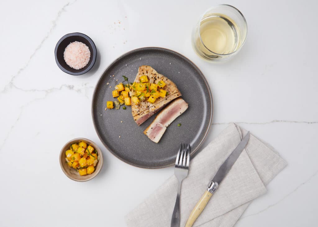 A beautifully plated dish featuring seared tuna steak topped with mango salsa, accompanied by a small bowl of diced mango, a pinch of pink salt, and a glass of white wine on a marble countertop.