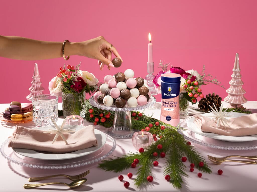 Hand reaching for chocolate truffle on a festive table set with macarons, flowers, and Morton Himalayan Pink Salt, accented with holiday decorations and a pink backdrop.