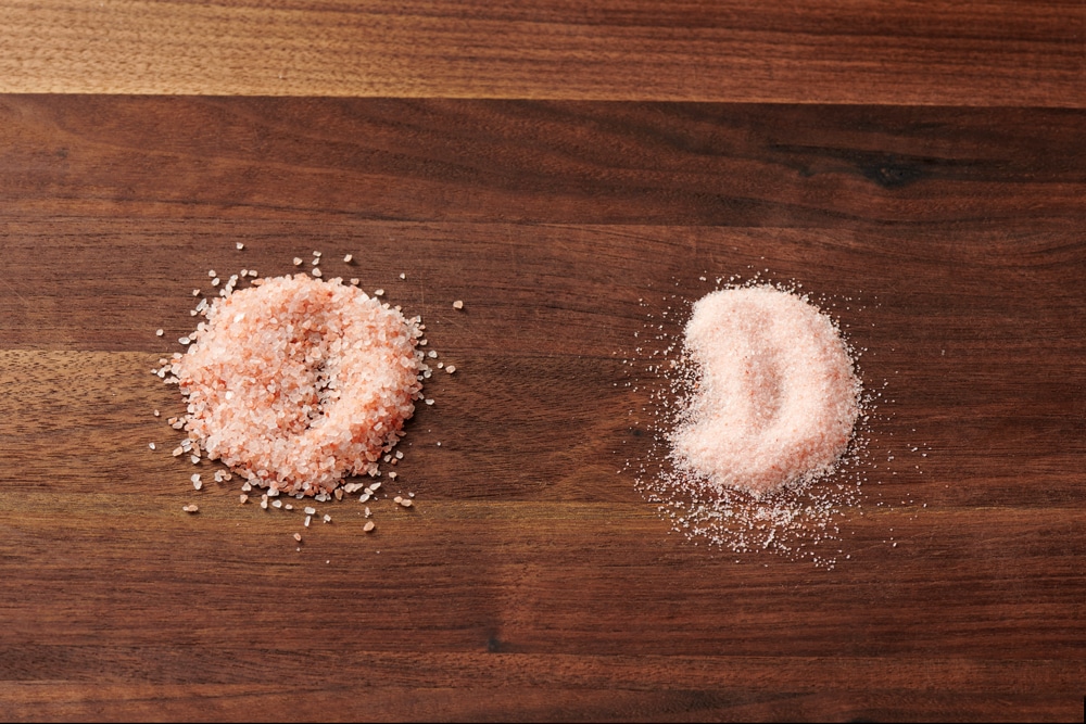 Close-up image of two types of pink salt on a wooden surface, showing coarse pink Himalayan salt on the left and fine pink salt on the right, highlighting their texture differences.