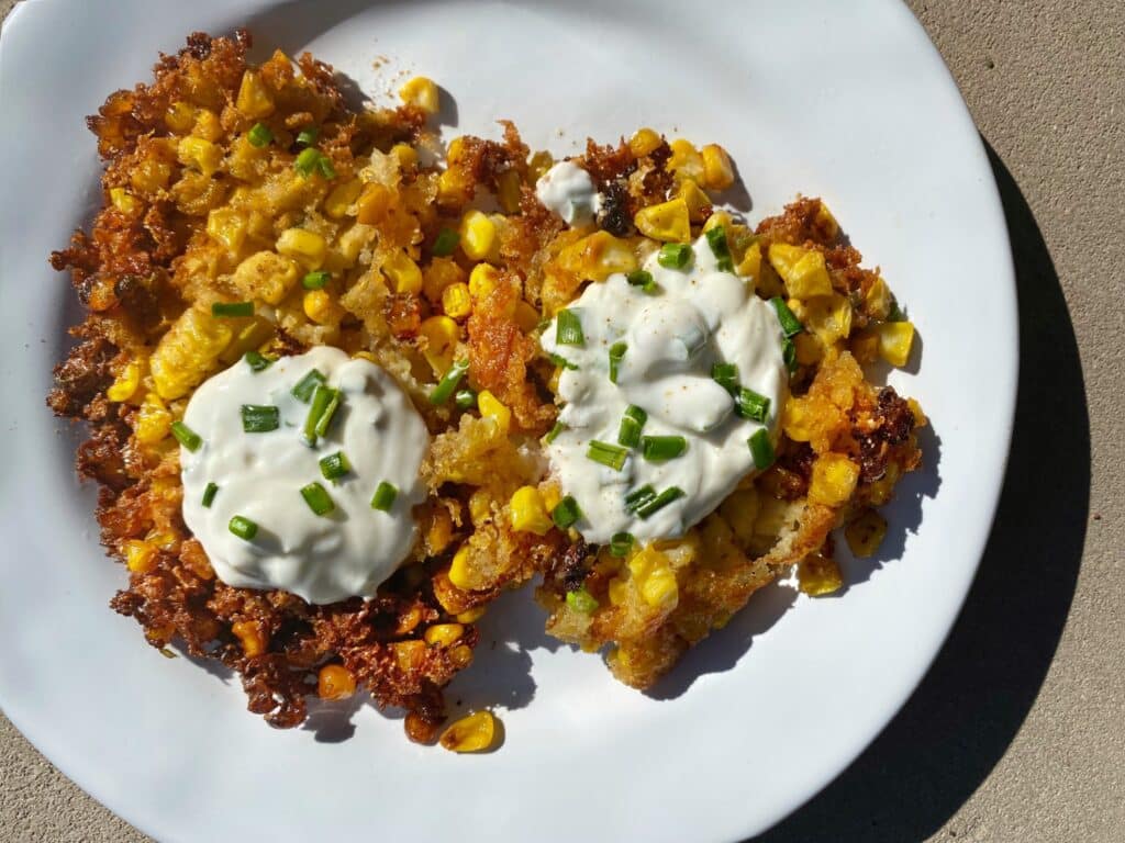 Golden corn fritters topped with sour cream and fresh chives, served on a white plate. The fritters are crispy on the outside and filled with sweet corn, highlighting a delicious, savory dish perfect for brunch or a snack.