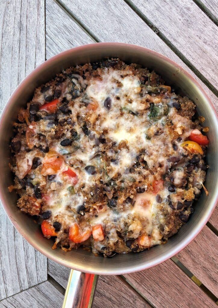 Baked quinoa dish with black beans, colorful bell peppers, and melted cheese, served in a metal pot on a wooden table. This healthy and nutritious meal is perfect for a quick dinner or meal prep.