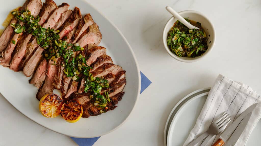 Sliced grilled steak topped with fresh herbs and a side of chimichurri sauce, served with grilled lemon halves on a white platter. A fork and knife are placed beside a clean plate and a linen napkin on a marble surface.