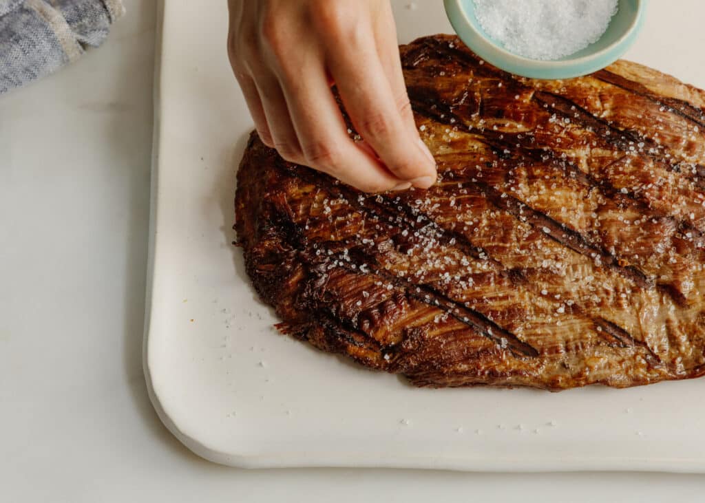 Hand sprinkling salt on a perfectly cooked steak resting on a white serving platter, showcasing its juicy texture and grill marks.