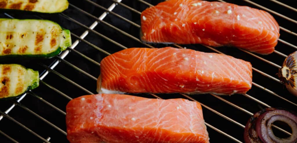 Grilled salmon fillets alongside zucchini and onion slices on a barbecue grill, showcasing a healthy and delicious cooking option.
