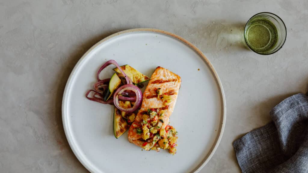 Grilled salmon fillet topped with a fresh salsa, served alongside sautéed zucchini and red onions on a white plate, with a glass of green drink and a gray linen napkin in the background.