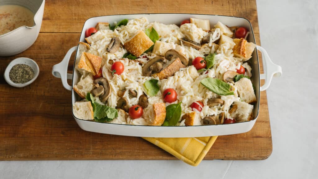 Baked casserole dish filled with bread cubes, cherry tomatoes, spinach, and sautéed mushrooms, topped with shredded cheese, served on a wooden cutting board with a side of seasoning and sauce.