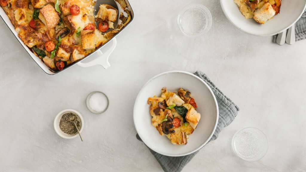 Baked vegetable casserole with tomatoes and mushrooms served in a white bowl, accompanied by a serving dish, salt, pepper, and sparkling water on a light-colored countertop.