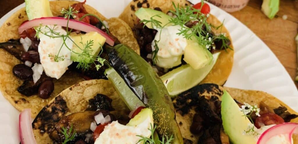 Delicious tacos topped with black beans, fresh avocado, lime wedges, pickled red onions, and garnished with cilantro, served on a paper plate.