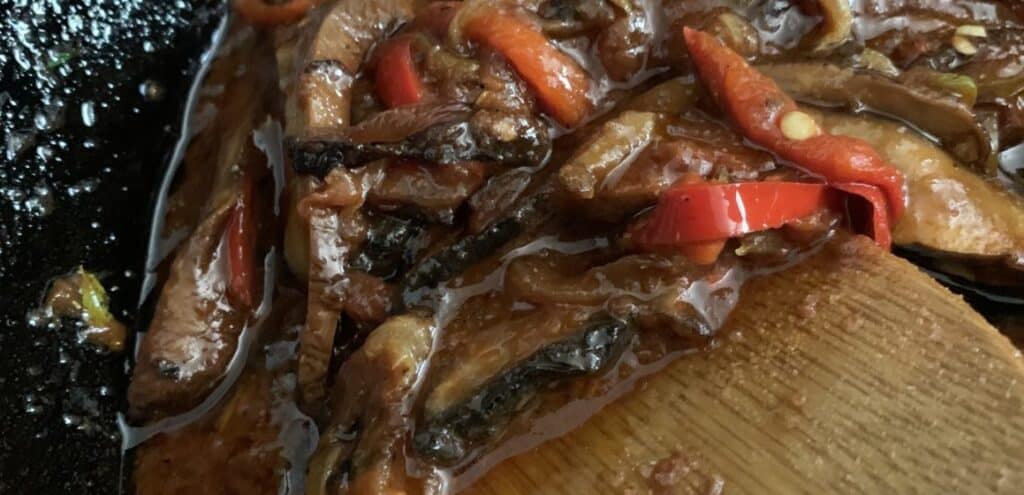Stir-fried mushrooms and red peppers in a glossy sauce, served in a black skillet with a wooden spatula, showcasing a vibrant and appetizing dish.